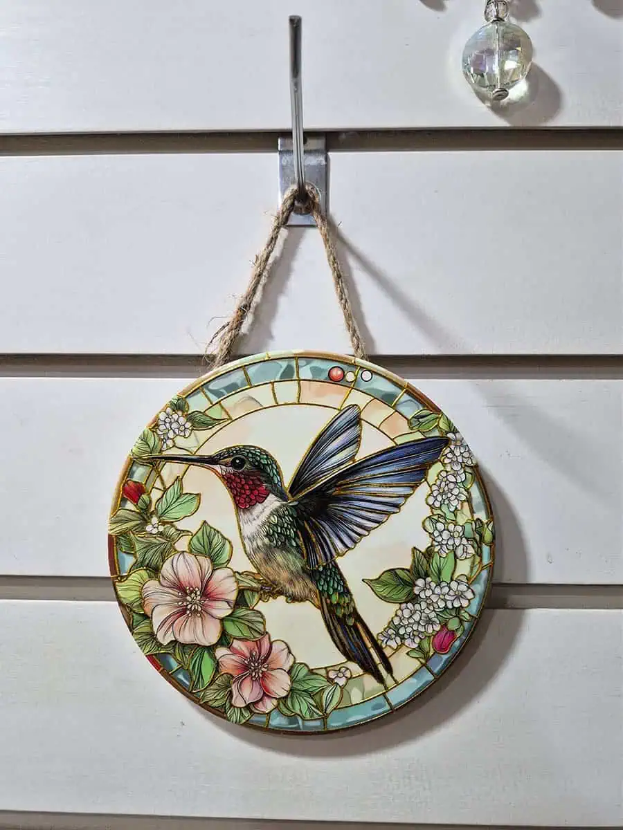 Colgante decorativo redondo de cerámica con colibrí entre flores blancas y rosadas en tonos celestes y verdes