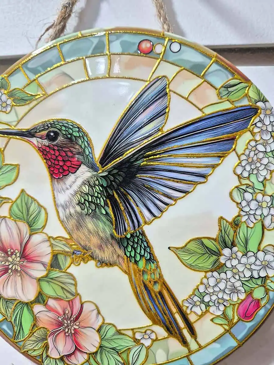 Colgante decorativo redondo de cerámica con colibrí entre flores blancas y rosadas en tonos celestes y verdes