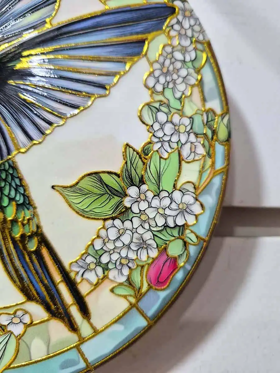 Colgante decorativo redondo de cerámica con colibrí entre flores blancas y rosadas en tonos celestes y verdes