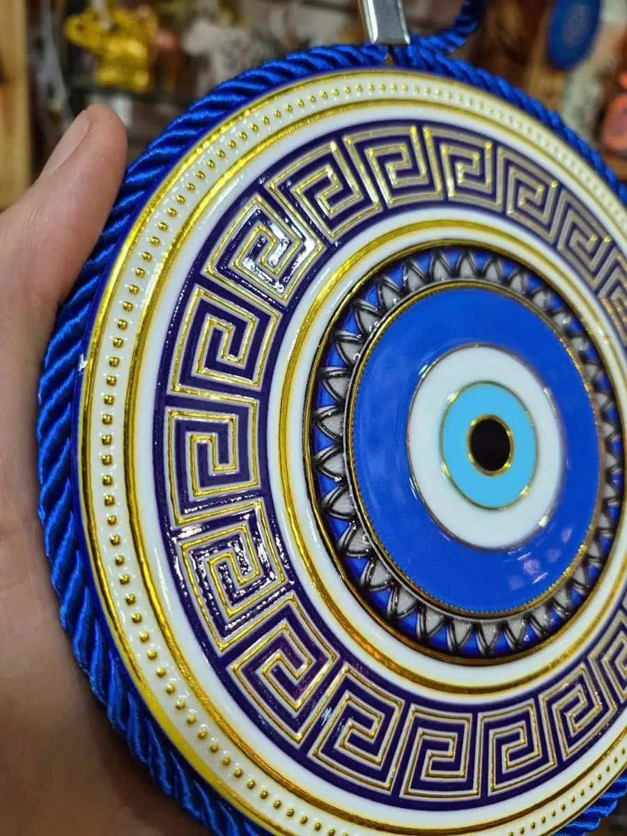 Colgante de ojo turco redondo azul y celeste con guarda griega azul oscuro, fondo blanco, detalles dorados y cordón azul
