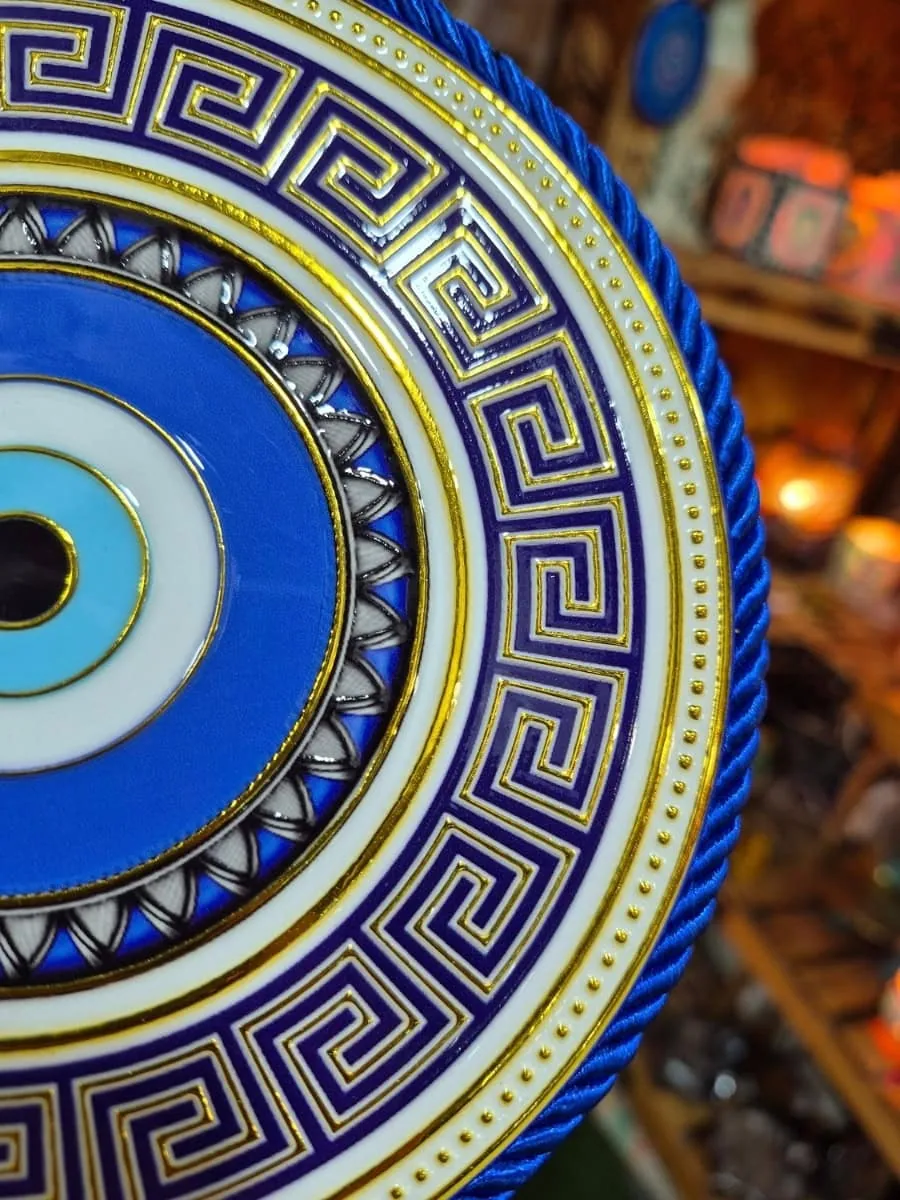 Colgante de ojo turco redondo azul y celeste con guarda griega azul oscuro, fondo blanco, detalles dorados y cordón azul