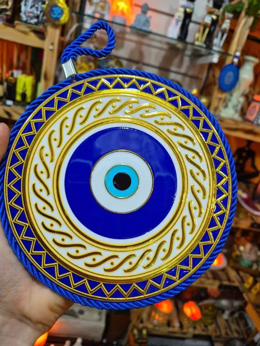 Ojo turco redondo azul con borde de triángulos dorados, dibujos curvos sobre fondo blanco y cordón trenzado azul