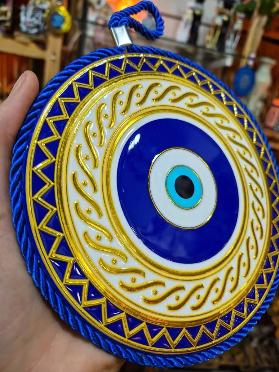 Ojo turco redondo azul con borde de triángulos dorados, dibujos curvos sobre fondo blanco y cordón trenzado azul