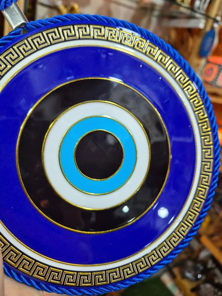 Colgante redondo de ojo turco azul con círculos negros, blancos y celestes, guarda griega dorada y cordón azul para colgar