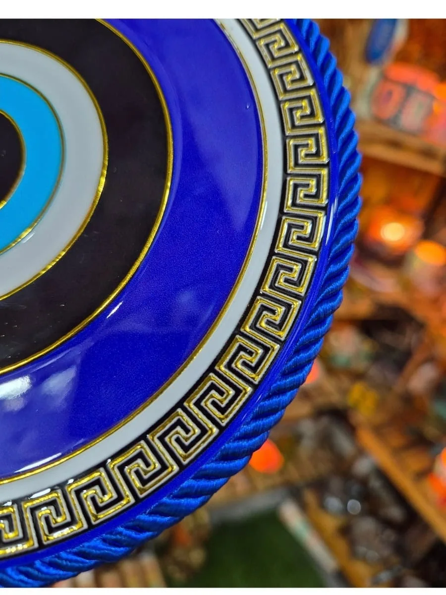 Colgante redondo de ojo turco azul con círculos negros, blancos y celestes, guarda griega dorada y cordón azul para colgar