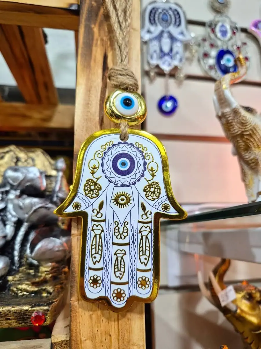 Mano de Fátima blanca con dibujos dorados, ojo turco azul central y detalle de ojo protector sobre cordón de soga