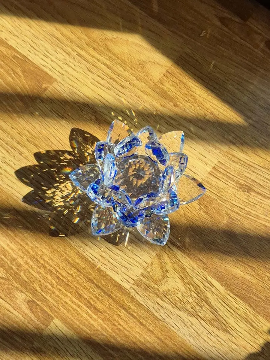 Flor de loto de vidrio transparente con detalles azules, pétalos facetados y destellos de luz solar sobre una superficie de madera