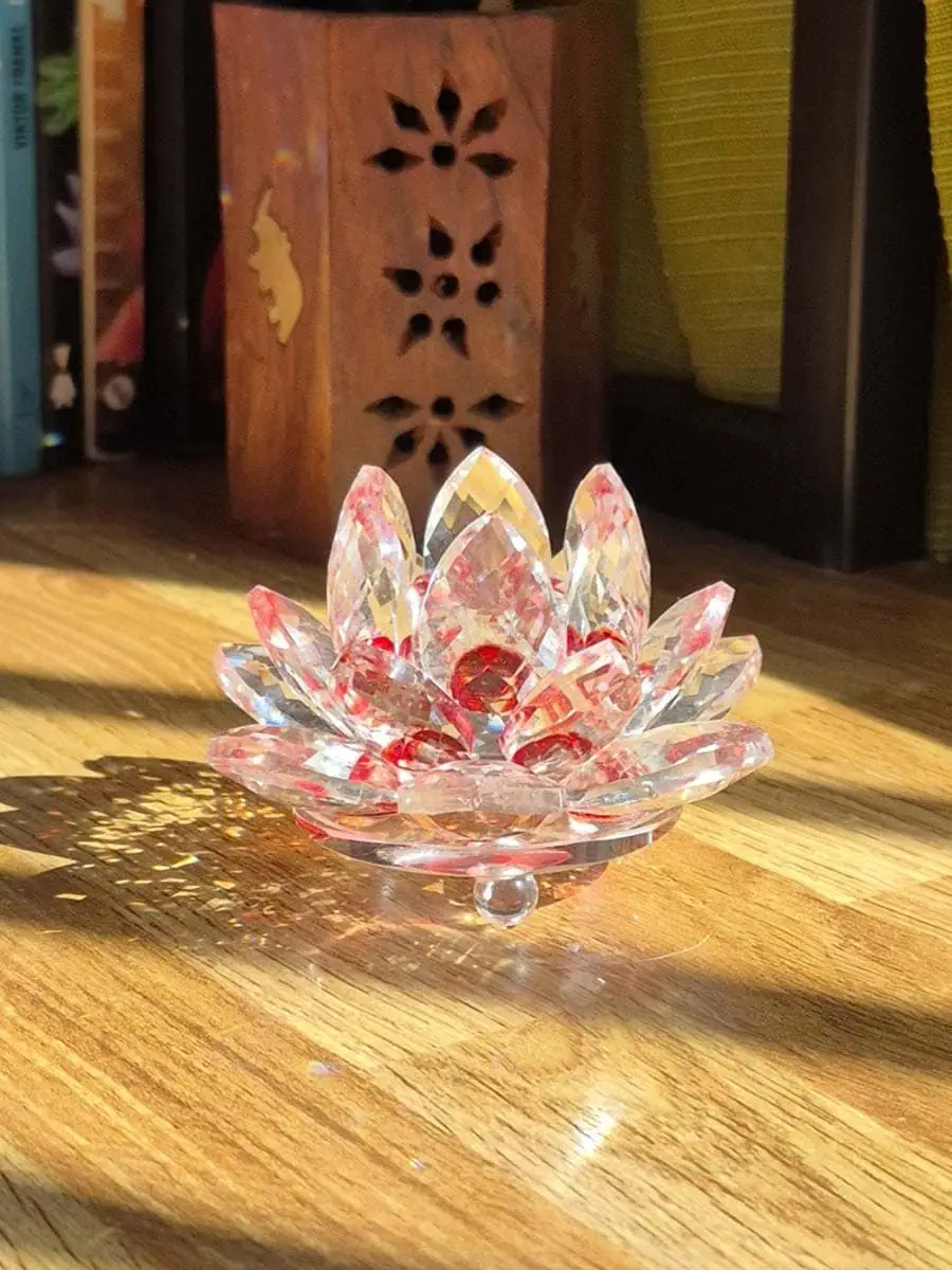 Flor de loto de vidrio transparente con detalles rojos y rosados, pétalos facetados que reflejan la luz del sol sobre madera