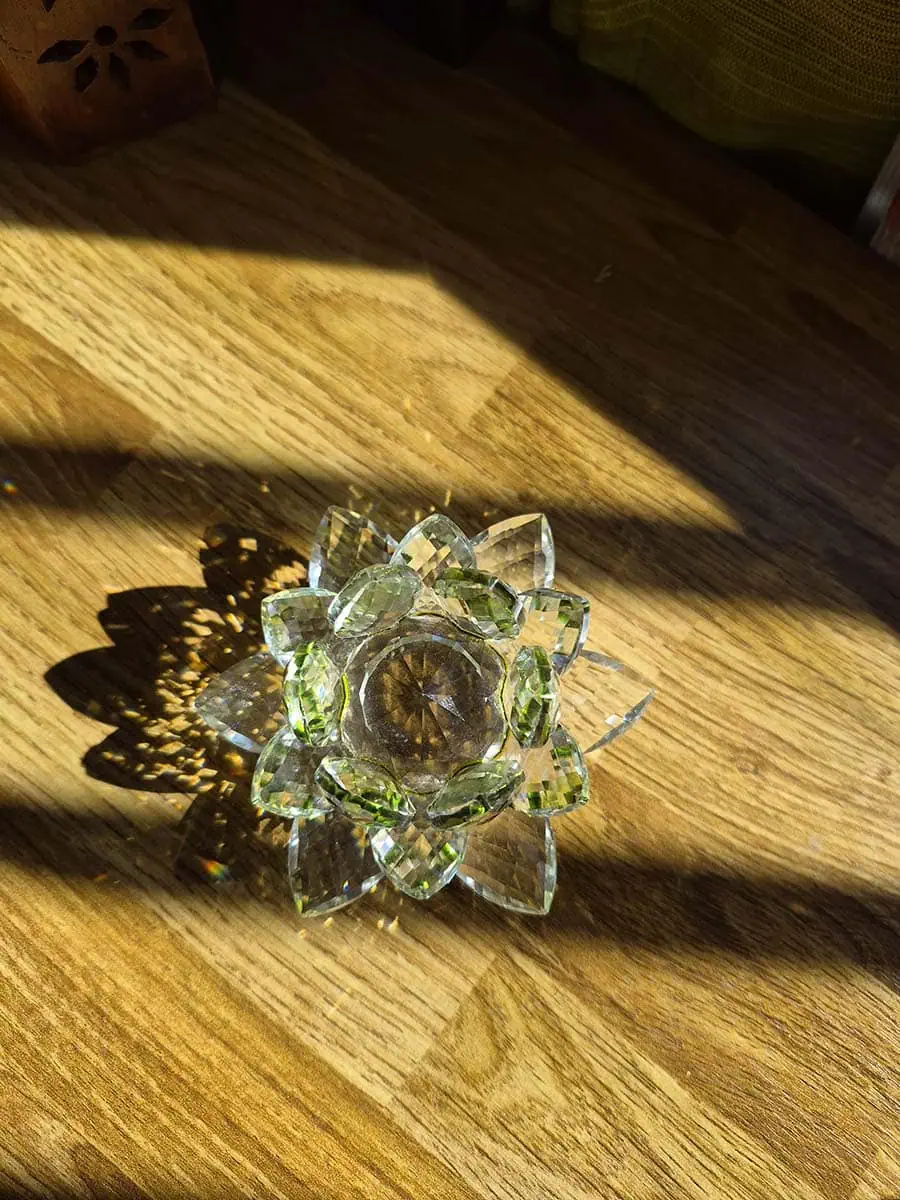 Flor de loto decorativa de vidrio transparente con detalles verdes, pétalos facetados y reflejos de luz solar sobre madera