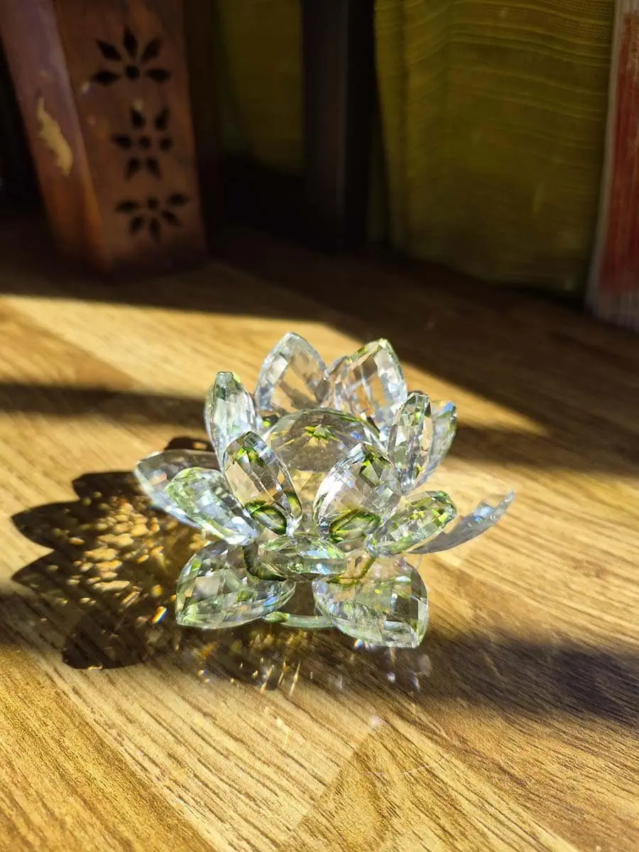 Flor de loto decorativa de vidrio transparente con detalles verdes, pétalos facetados y reflejos de luz solar sobre madera